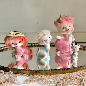 Pode incluir: Três estatuetas de cerâmica de um gato rosa, um cão rosa com chapéu e um gato branco. As estatuetas estão sobre uma bandeja dourada com uma superfície espelhada.