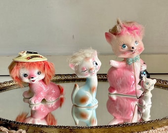 以下が含まれることがあります： ピンクの猫、帽子をかぶったピンクの犬、白い猫のセラミック製置物3つ。置物は鏡面仕上げの金色のトレイの上に置かれています。