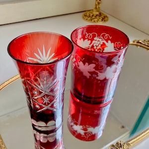 May include: Two red glass shot glasses with etched white designs. One glass has a starburst pattern, the other has a grapevine pattern.