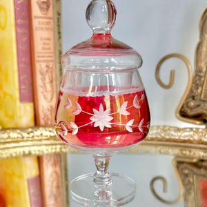 Op de afbeelding: Een heldere glazen pot met een deksel, met een levendig rood en goud interieur. De pot is versierd met witte bloemen- en bladmotieven. Hij staat op een heldere, voetvormige basis, die het licht reflecteert. De pot is waarschijnlijk een decoratief stuk.