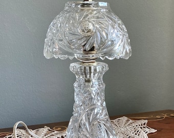 May include: A clear crystal table lamp with a cut glass shade and a silver finial. The lamp has a white cord and is sitting on a crocheted doily.