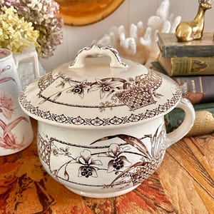 May include: A cream-colored ceramic chamber pot with a lid, featuring a brown transferware pattern of flowers, leaves, and woven basket motifs. The piece includes a curved handle on the side and a decorative knob on the center of the lid.