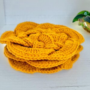 May include: A stack of handmade, mustard-yellow crochet coasters. The coasters are circular with a textured, floral design. The yarn appears thick and soft, with visible stitch detail. The coasters are stacked on a white surface.