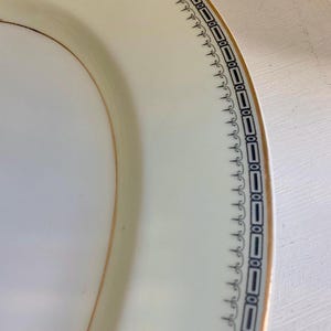May include: Cream-colored oval serving platter with a gold rim and a decorative border. The border features a repeating pattern of black rectangles and scrollwork. The plate is made of porcelain.