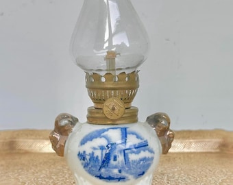May include: A small, vintage oil lamp with a clear glass chimney and brass accents. The white ceramic base features a blue and white windmill scene. The lamp has a decorative design and is likely a collectible item.
