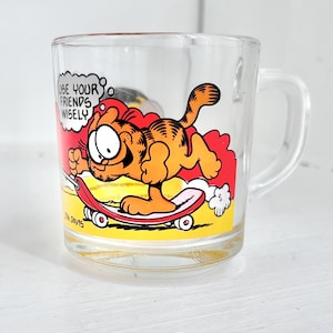 Może przedstawiać: Przezroczysty szklany kubek z uchwytem, przedstawiający kota Garfielda na deskorolce. Kubek ma kolorową grafikę Garfielda na deskorolce z tekstem "USE YOUR FRIENDS WISELY" w dymku.