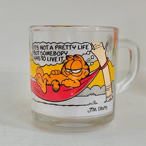 Może przedstawiać: Przezroczysty szklany kubek z Garfieldem w hamaku z cytatem "IT'S NOT A PRETTY LIFE BUT SOMEBODY HAS TO LIVE IT." Kubek ma uchwyt i kolorowy design z pomarańczowymi, czerwonymi, żółtymi i białymi akcentami. Podpisano "JPM DAVIS".