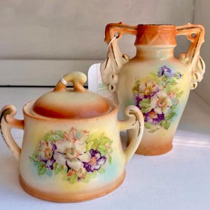 May include: A vintage porcelain sugar bowl and creamer set with floral designs. The sugar bowl has a lid and both pieces have gold accents. The floral designs are in shades of purple, white, and yellow.