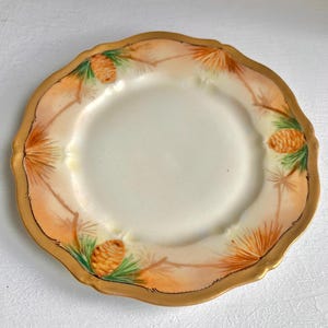 May include: A white porcelain plate with a gold rim and a delicate design of pine branches and pine cones in shades of orange, brown, and green.