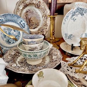 May include: A collection of vintage ceramic tableware including several patterned plates, stacked bowls, and teacups with saucers. The items feature blue, brown, and green transferware designs. A brass candlestick and a brass horse figurine are present.