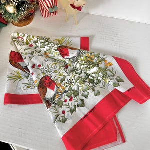 May include: A white kitchen towel featuring a repeating pattern of red-breasted robins, green ivy leaves, and red berries. The towel has a solid red border along the edges and includes the printed text LAMONT reg1803 and MADE IN.