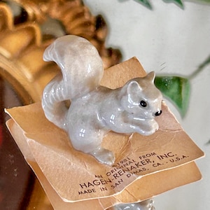 May include: A small, ceramic squirrel figurine in shades of gray, with a fluffy tail and black eyes. The squirrel is posed on a small, folded paper tag with text that reads "HAGEN-RENAKER, INC. MADE IN SAN DIMAS, CA. U.S.A."