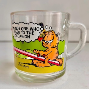 Może przedstawiać: Przezroczysty kubek ze szkła z kreskówkowym wizerunkiem Garfielda kota na huśtawce. Kubek ma uchwyt i napis "I'm not one who rises to the occasion" w dymku nad Garfieldem. Na spodzie kubka widnieje napis "Jim Davis".