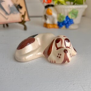 May include: A ceramic figurine of a brown and white dog lying down with its head resting on its paws. The dog has brown eyes and a brown nose. The figurine is painted with brown and white details.