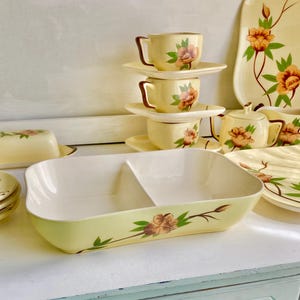 Puede incluir: Un juego de vajilla de cerámica vintage con estampado floral amarillo y marrón, que incluye una fuente de servir dividida, tazas de té y platillos, una jarra de crema y un azucarero.