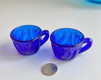 May include: Two cobalt blue glass teacups with a floral design. The cups have a small handle and are about 3 inches tall.