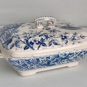 May include: A white ceramic serving dish with a blue and white floral pattern. The dish has a lid with a handle and a square base.