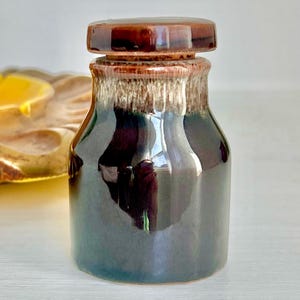 May include: A small, dark brown ceramic jar with a matching lid. The jar has a glossy finish and a textured, light brown band around the neck. The lid is flat and round, and the jar appears to be empty.