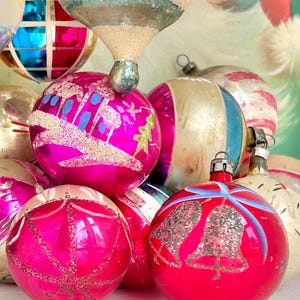 May include: A collection of vintage Christmas ornaments in various shapes and colors. The ornaments are primarily pink, red, and silver, with some featuring painted designs and glitter accents. The collection includes round and teardrop-shaped ornaments.