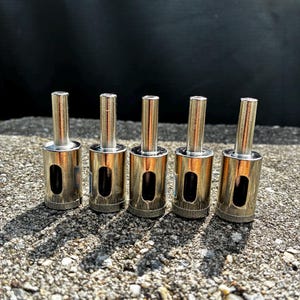 May include: Five silver-colored metal diamond hole saw drill bits arranged in a row. Each tool features a cylindrical shank, a hollow barrel body with a rectangular side opening, and a textured abrasive cutting edge at the base.