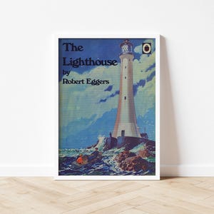 May include: Framed art print depicting a lighthouse on a rugged coastline. The image shows a tall, white lighthouse against a backdrop of a blue sky with clouds. The title "The Lighthouse" by Robert Eggers is visible at the top.