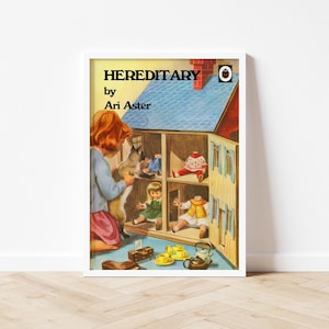May include: A framed print featuring the book cover of "Hereditary" by Ari Aster. The artwork depicts a young girl with red hair interacting with a dollhouse containing dolls. The dollhouse has a blue roof and yellow walls. The title and author's name are in black text.