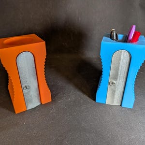 Puede incluir: Dos coloridos organizadores de escritorio para portalápices con forma de sacapuntas gigantes. Uno es naranja, el otro azul, ambos con un diseño de cuchilla plateada. El azul contiene bolígrafos y lápices.