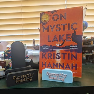 May include: A paperback book titled On Mystic Lake by Kristin Hannah with an orange cover, displayed behind two book stands. One stand is black with gold text reading Currently Reading, and the other is light blue with white text reading Currently Reading.