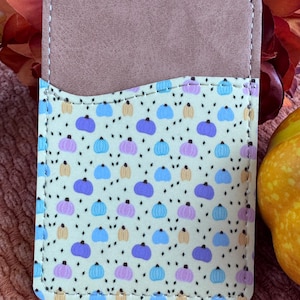 May include: A rectangular phone card holder with a light brown top and a patterned front. The front features a repeating pattern of blue, purple, and yellow pumpkins on a cream background, with black diamond accents. The edges are stitched with white thread.