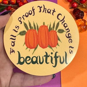 May include: A round, yellow sticker with the text "Fall is proof that change is beautiful" in dark brown lettering. Three orange pumpkins with green leaves are illustrated in the center of the sticker.