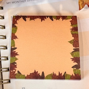 May include: Square sticky note pad with a border of autumn leaves in shades of brown, green, and orange. The center of the pad is a solid peach color, providing space for notes. The pad is resting on a spiral-bound notebook.