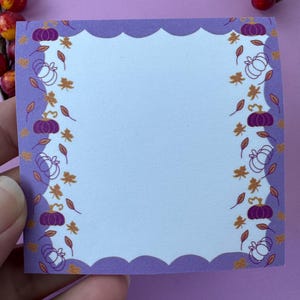 May include: A square, light blue notepad with a scalloped purple border. The border features a repeating pattern of purple and white pumpkins, and orange leaves. The notepad is held by a hand, with a purple background.