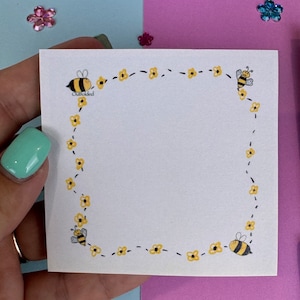 May include: A white square sticky note with a bee and flower border. The note has the word "Outfolded" written on it.