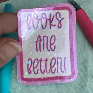 May include: A rectangular sticker with a pink border and the words "books are better!" in purple script. The background has a sparkly, iridescent effect with small star shapes. A blue pen and a pink pen are visible.