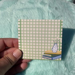 May include: Square sticky note with a green grid pattern. The design features a stack of books and a vase with white flowers. The note is held against a light blue, plush background.