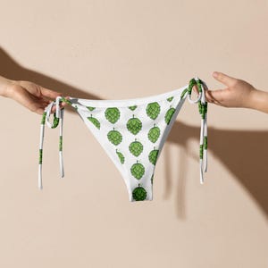 May include: White bikini bottoms with a repeating pattern of green hops. The bikini bottoms have white ties with green accents. The garment is held up by two hands against a neutral background.