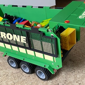 Può includere: Un rimorchio giocattolo verde con una griglia nera e la scritta "KRONE" stampata sul lato. Il rimorchio è pieno di blocchi di giocattolo colorati e ha uno sportello posteriore incernierato che è aperto.