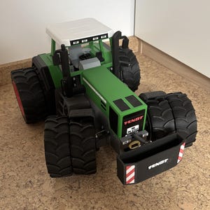 May include: A green and grey toy tractor with large black tyres and a white roof. The tractor has the word "FENDT" printed on the front and a black front attachment with red and white stripes.