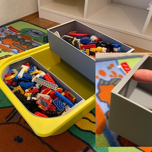 Op de afbeelding: Een gele plastic opbergbak met een grijs deksel gevuld met kleurrijke Lego-blokken. De bak staat op een kleurrijk tapijt. Er is een grijze opbergbak op de achtergrond.