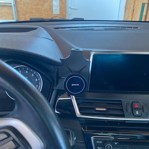 Puede incluir: Salpicadero negro de coche con soporte para teléfono. El soporte sujeta un dispositivo circular negro con el texto "lomicoll". El salpicadero incluye una pantalla digital, volante y varios controles.