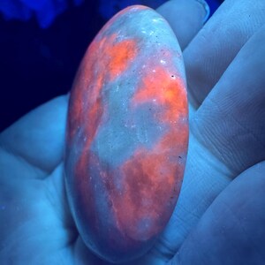 May include: An oval-shaped, polished stone with a glowing, fiery orange pattern against a pale, translucent background. The stone is held in a hand, with the background illuminated by a blue light.