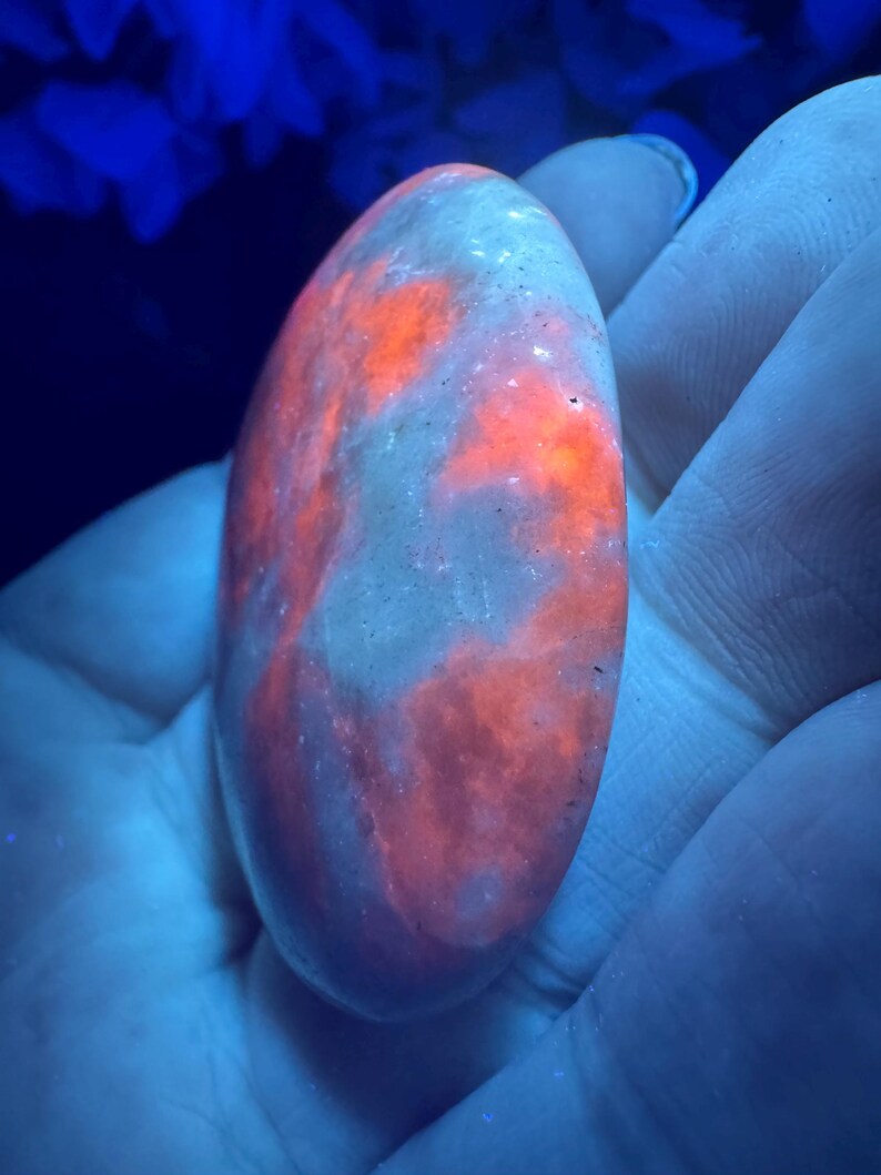 May include: An oval-shaped, polished stone with a glowing, fiery orange pattern against a pale, translucent background. The stone is held in a hand, with the background illuminated by a blue light.