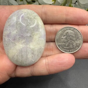 May include: An oval-shaped, polished stone with a mix of lavender and cream colors. The stone is smooth and has a natural, organic appearance. A US quarter coin is next to the stone for size comparison.