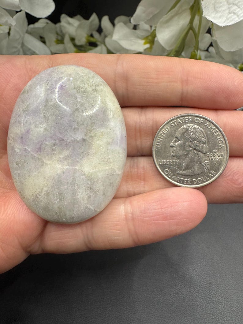 May include: An oval-shaped, polished stone with a mix of lavender and cream colors. The stone is smooth and has a natural, organic appearance. A US quarter coin is next to the stone for size comparison.