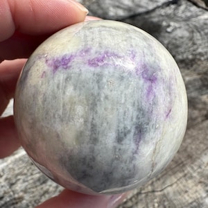 May include: A polished, spherical stone with a mix of colors. The stone features a base of light gray and white, with streaks of purple running across its surface. The sphere is held in a hand.