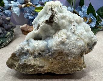 May include: A large, white and gray, geode with a cluster of small, round, blue-gray crystals. The geode is sitting on a light gray surface.