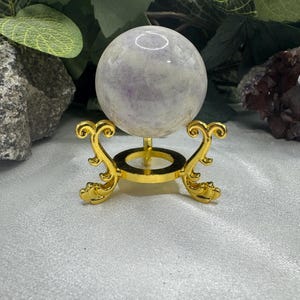 May include: A light purple crystal sphere displayed on a gold-colored metal stand. The stand has an ornate, scrolled design. The sphere has a translucent appearance with subtle color variations. The background includes greenery and rocks.