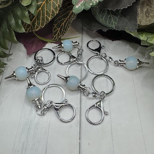 May include: A set of six silver keychains with a clear, iridescent, round, crystal-like charm attached to each. The charms are attached to the keychains with a silver spike.