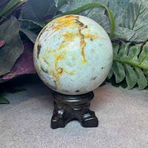 May include: A polished, spherical stone with a smooth, off-white base color featuring prominent, irregular veins of golden-yellow and dark brown mineral inclusions. The sphere rests securely on a dark, carved wooden tripod display stand.