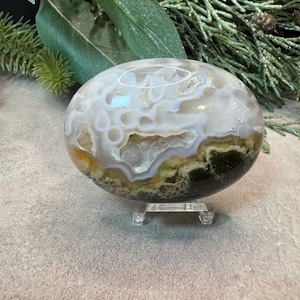 May include: A polished, oval-shaped agate stone featuring translucent white and grey swirling patterns with a central crystalline geode cavity. The stone displays deep green and earthy brown mineral inclusions at the base and rests on a clear stand.